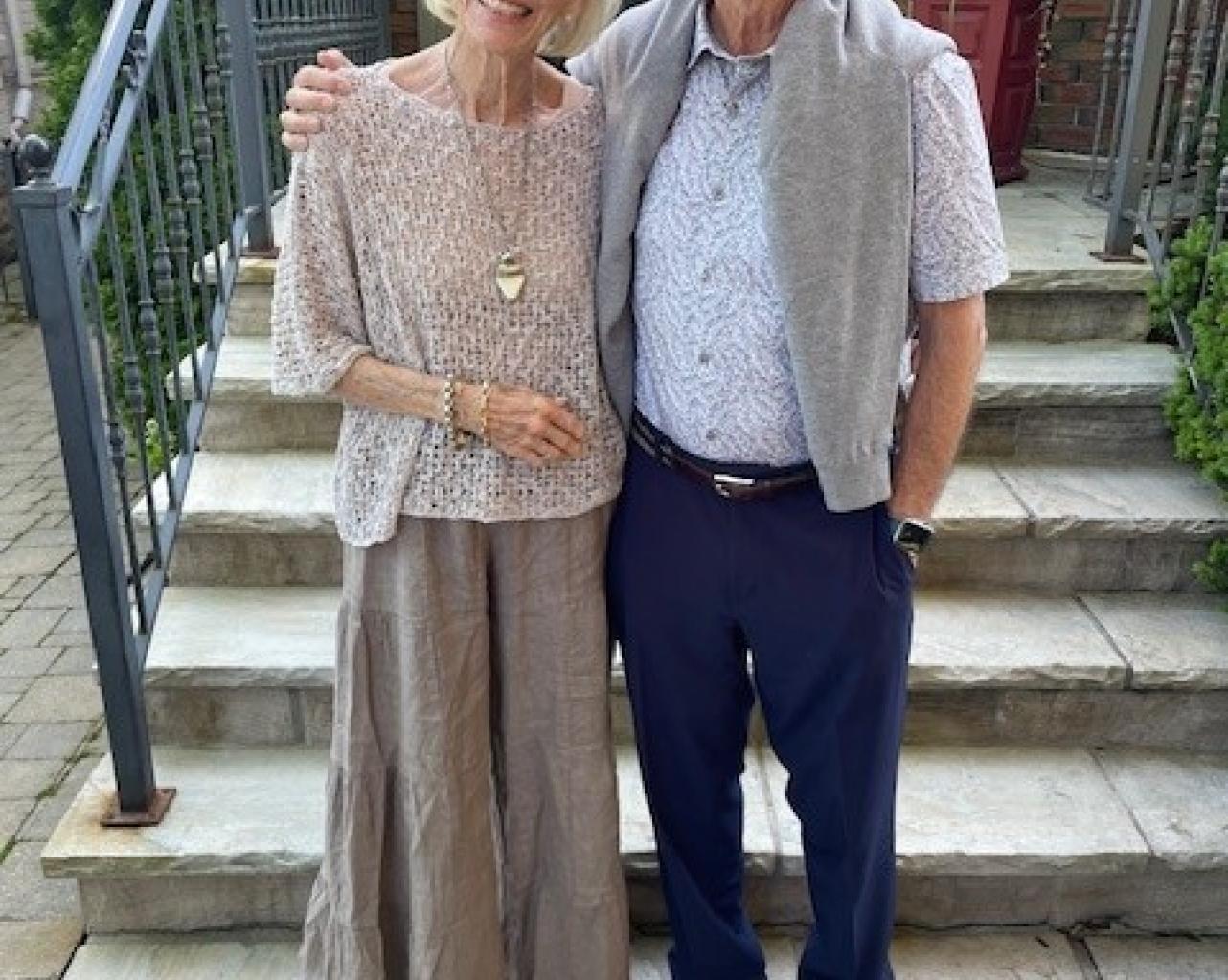 Jan and Tom, smiling in front of some steps outside. Jan is wearing a tan coloured shirt and skirt and Tom is wearing a light blue shirt, grey sweater around his shoulders, and blue pants..