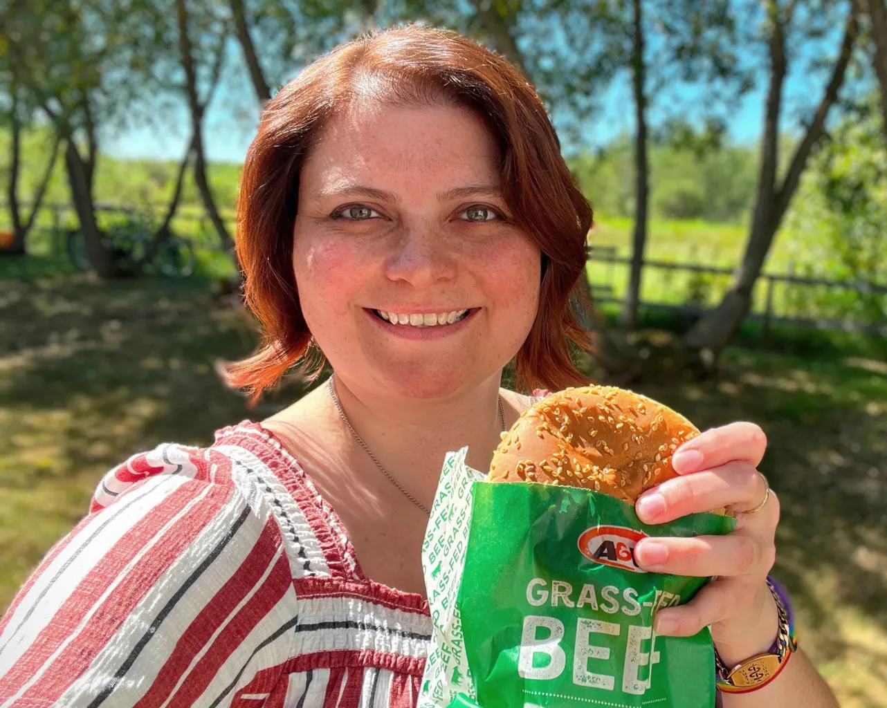 Caitlin sourit en tenant un Teen Burger™. Elle est à l’extérieur. En arrière-plan apparaissent des arbres.