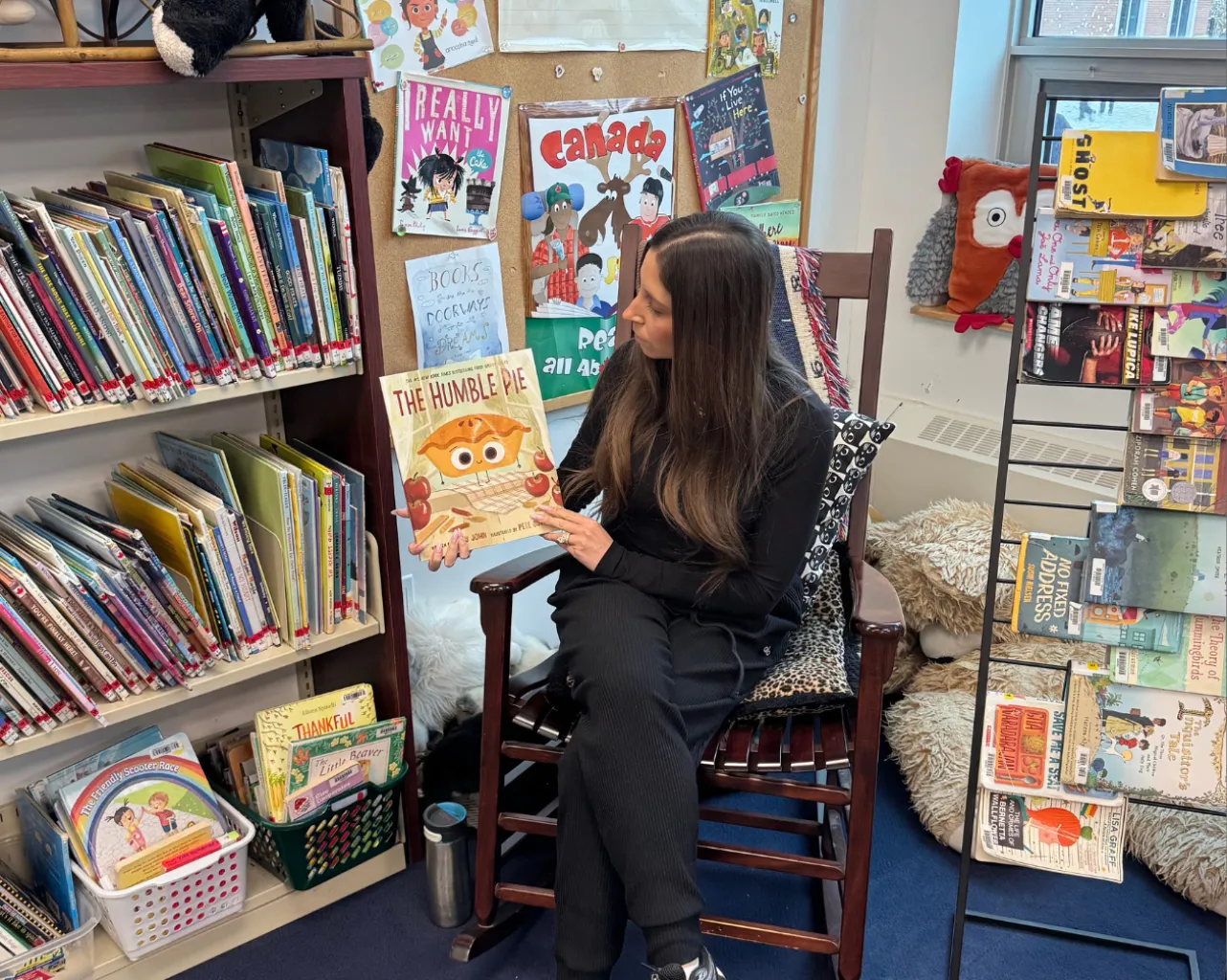 Jaclyn, wearing a black top and black pants, sitting in a chair, holding up a book.