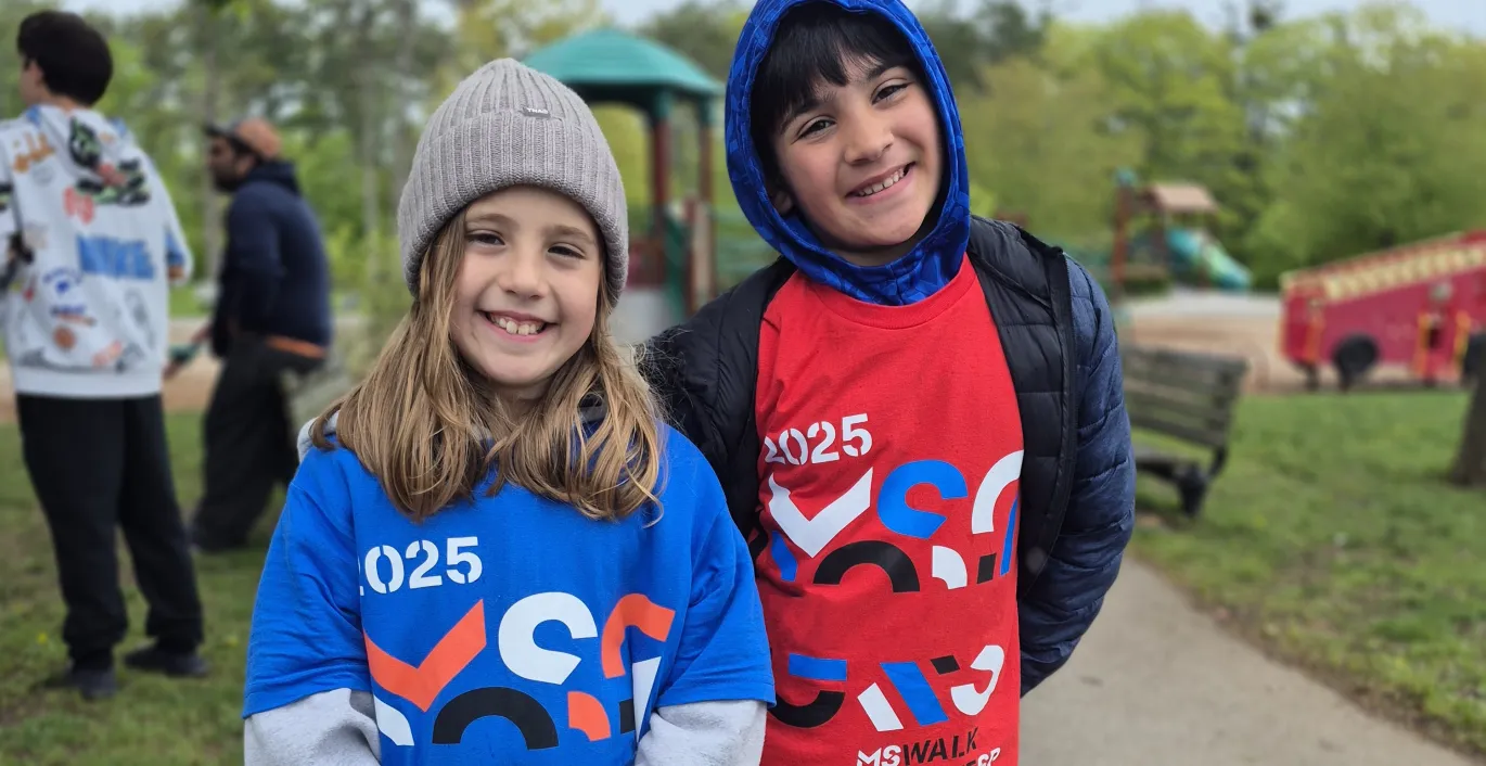 Two smiling children wearing numbered race bibs outdoors at a park event.