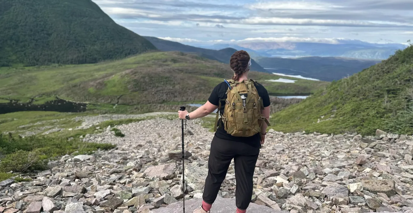 Janice, équipée d’un sac à dos jaune, se tient sur un sentier de montagne rocailleux surplombant une vallée pittoresque.