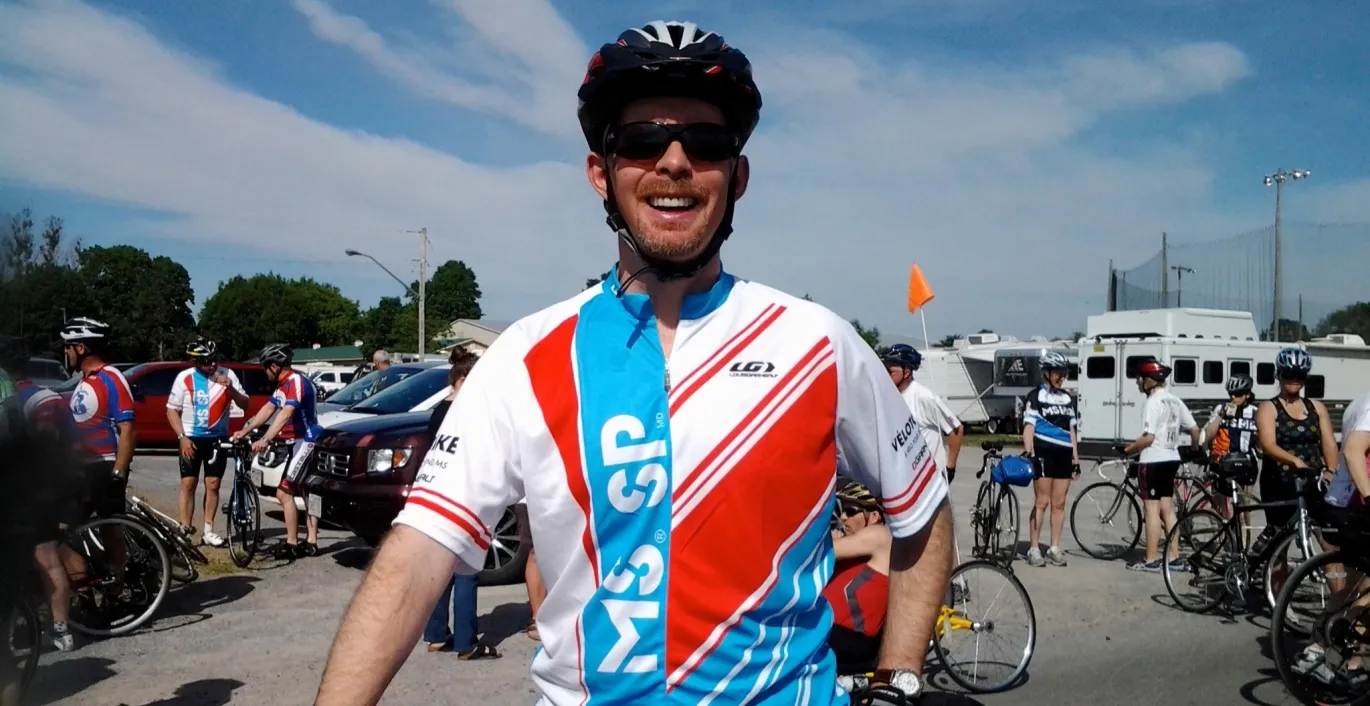 Craig porte un maillot du Vélo SP. Il est à l’extérieur et se tient devant son vélo.