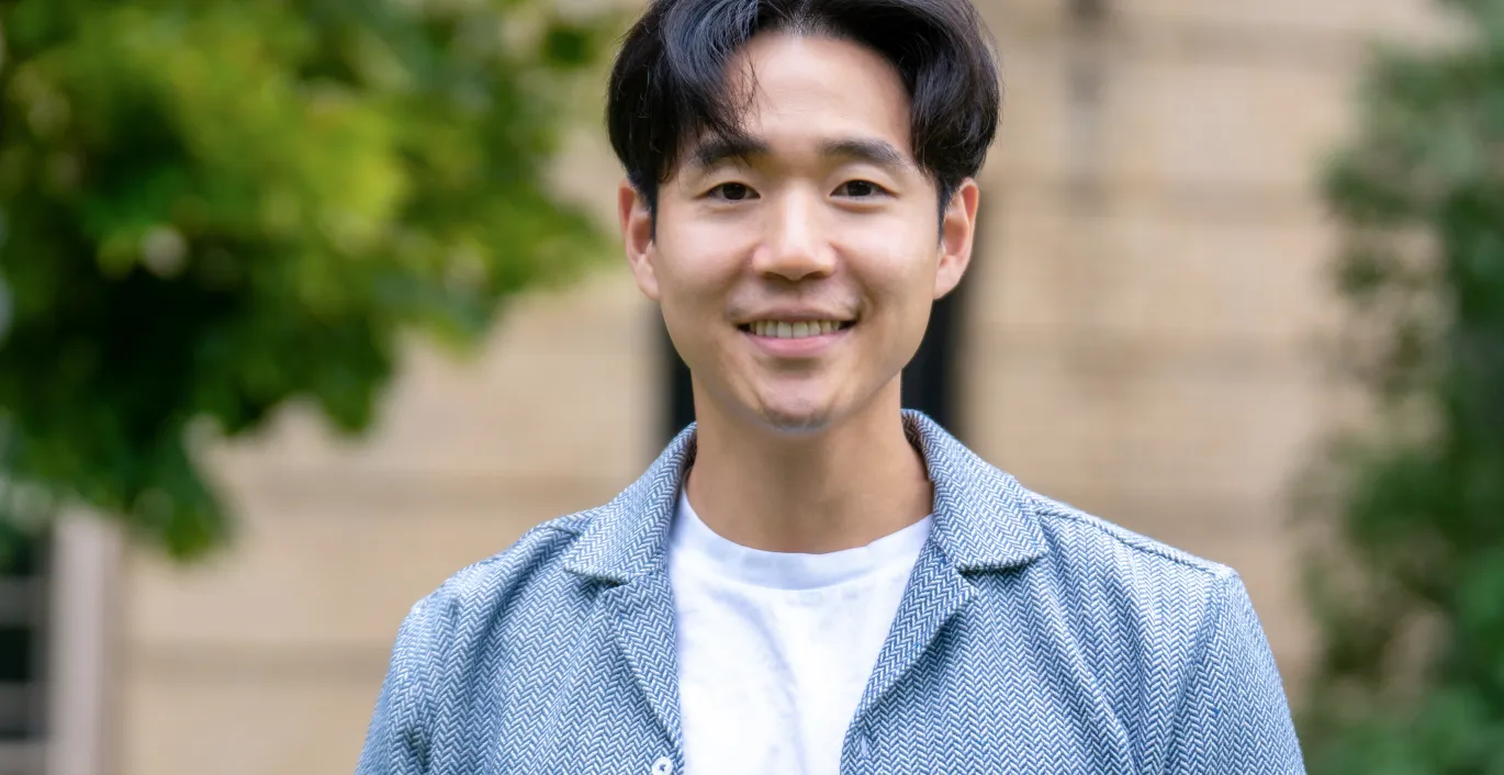 Paul Yoo, Ph. D., porte un t-shirt blanc et une veste bleu pâle. Il sourit et se tient debout devant un immeuble bordé d’arbres.