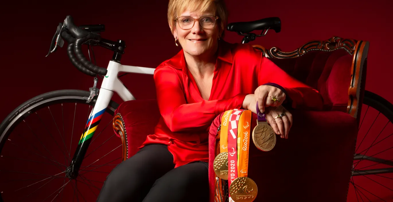 Carol porte un chemisier rouge et un pantalon noir. Elle est assise dans un fauteuil rouge devant un vélo de route et elle tient des médailles.