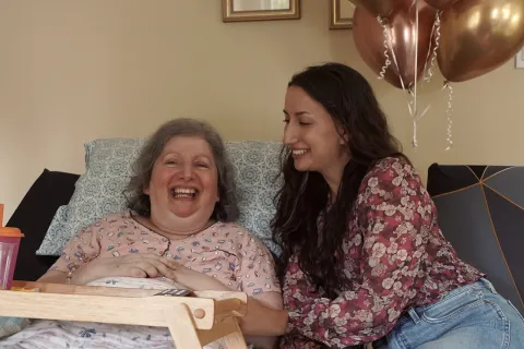 Stephanie est vêtue d’une blouse à motif fleuri. Elle sourit à sa mère, Carm, qui est allongée dans son lit et qui porte un pyjama rose. Des ballons roses apparaissent en arrière-plan.