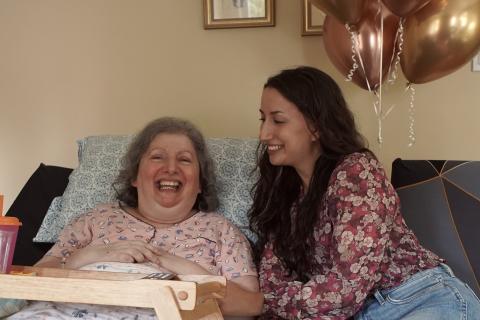 Stephanie est vêtue d’une blouse à motif fleuri. Elle sourit à sa mère, Carm, qui est allongée dans son lit et qui porte un pyjama rose. Des ballons roses apparaissent en arrière-plan.