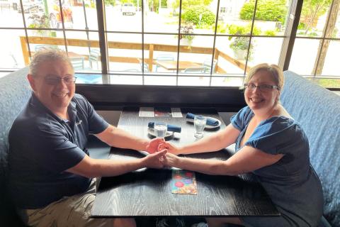 James, wearing a black shirt, and Yolanda, wearing a blue dress, sitting at a restaurant booth and holding hands.