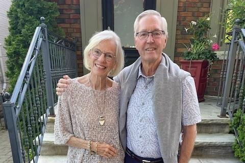 Jan and Tom, smiling in front of some steps outside. Jan is wearing a tan coloured shirt and skirt and Tom is wearing a light blue shirt, grey sweater around his shoulders, and blue pants..