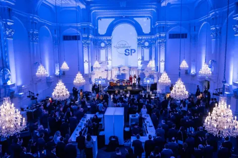 Elegant evening scene at the 2024 Cocktail Signature SP, with guests gathered in a grand hall under crystal chandeliers, facing a stage with speakers.