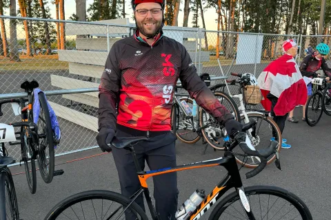 Brennan sourit et se tient debout, devant son vélo. Il porte un casque de vélo, des lunettes, des gants noirs, un pantalon noir, ainsi qu’une veste noir et rouge. En arrière-plan se trouvent d’autres vélos placés contre une clôture métallique.