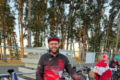 Brennan sourit et se tient debout, devant son vélo. Il porte un casque de vélo, des lunettes, des gants noirs, un pantalon noir, ainsi qu’une veste noir et rouge. En arrière-plan se trouvent d’autres vélos placés contre une clôture métallique.