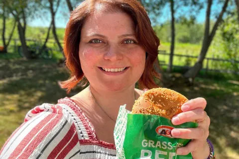 Caitlin sourit en tenant un Teen Burger™. Elle est à l’extérieur. En arrière-plan apparaissent des arbres.