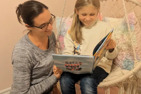 Izzy, wearing a beige sweater and jeans, sitting on a swing while holding a book beside her mother, Lindsey, wearing a grey top and jeans.