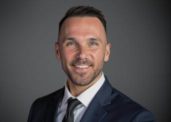 A man with short dark hair, a beard and blue eyes is wearing a navy coloured suit.