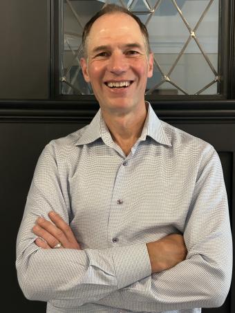 A man with short, brown hair, smiling broadly. He wears a light patterned collared shirt with crossed arms