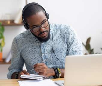 A man with very short brown hair and glasses is wearing headphones, a blue shirt and writing notes while viewing a laptop.