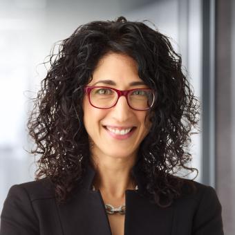 A woman with curly dark hair and red glasses, smiling and wearing a black blazer and chain-link necklace.