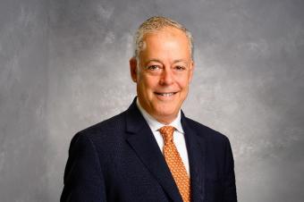 A man with short light coloured hair is wearing a black suit with an orange tie and white shirt.