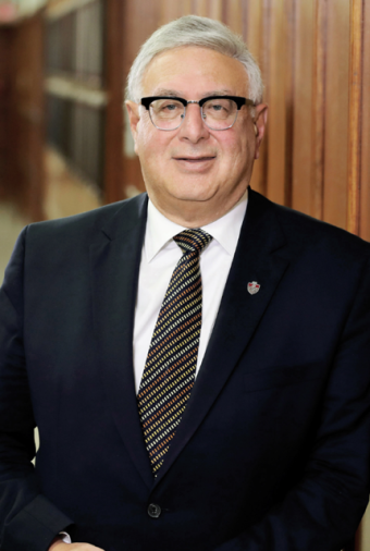 A man with white hair and black rimmed glasses is wearing a black suit with a diagonal striped tie.