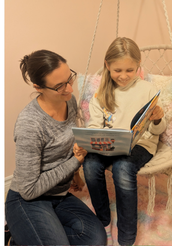 Izzy, wearing a beige sweater and jeans, sitting on a swing while holding a book beside her mother, Lindsey, wearing a grey top and jeans.