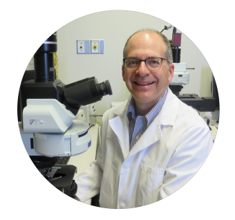 Smiling man in white lab coat and glasses standing next to a microscope in a laboratory.