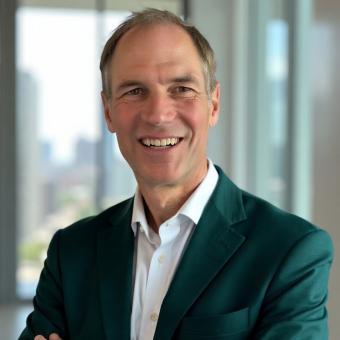 Smiling middle-aged man in a teal blazer with a city skyline visible in the background.