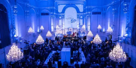 Elegant evening scene at the 2024 Cocktail Signature SP, with guests gathered in a grand hall under crystal chandeliers, facing a stage with speakers.