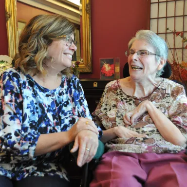 Two women smiling and chatting warmly together indoors, one younger and one elderly.