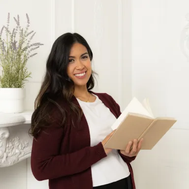 Smiling young woman with long dark hair holding an open book, standing near a white fireplace mantel.