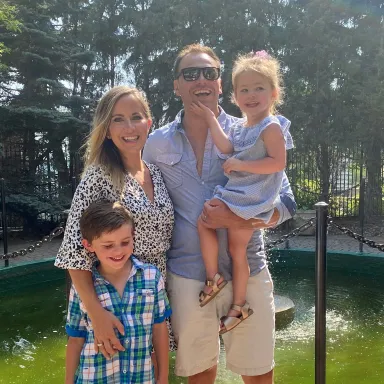 Family of four smiling outdoors on a sunny day, with two adults and two young children.