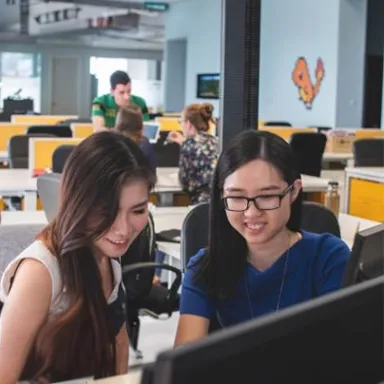 Two young women smiling while collaborating at a computer in a modern open office.