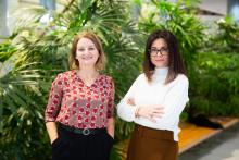 Two smiling women standing together in front of lush green tropical plants.
