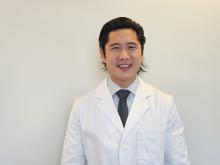 Smiling man in a white lab coat and dark tie against a light gray background.