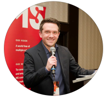 Man in suit speaking into a microphone at a conference, standing near a red banner.