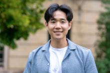Smiling young man in a light blue shirt outdoors with blurred greenery background.