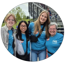 Four women smiling outdoors at an MS charity event, wearing teal MS volunteer shirts.