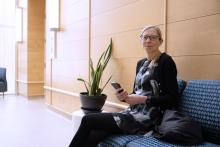 Woman in glasses sitting in a modern lobby, holding a phone, with a green plant beside her.