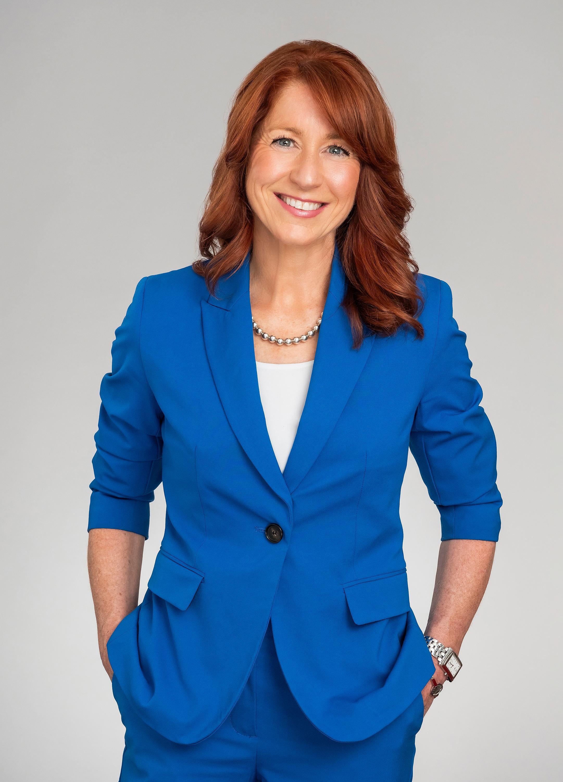 A woman with red hair smiling and wearing a blue blazer, with her hands in her pockets.