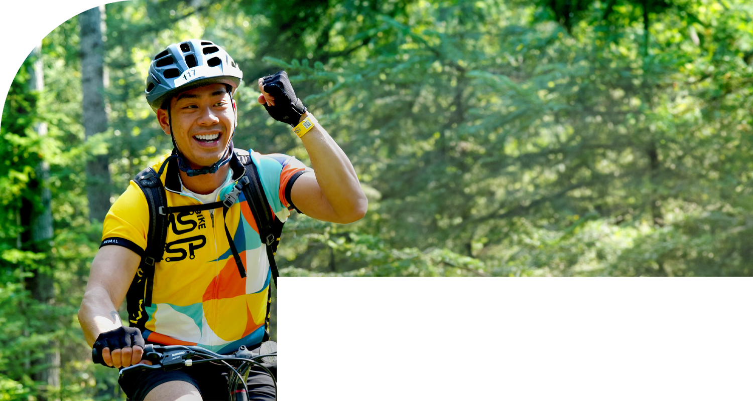 A bike rider riding near trees while smiling and pumping his arm.