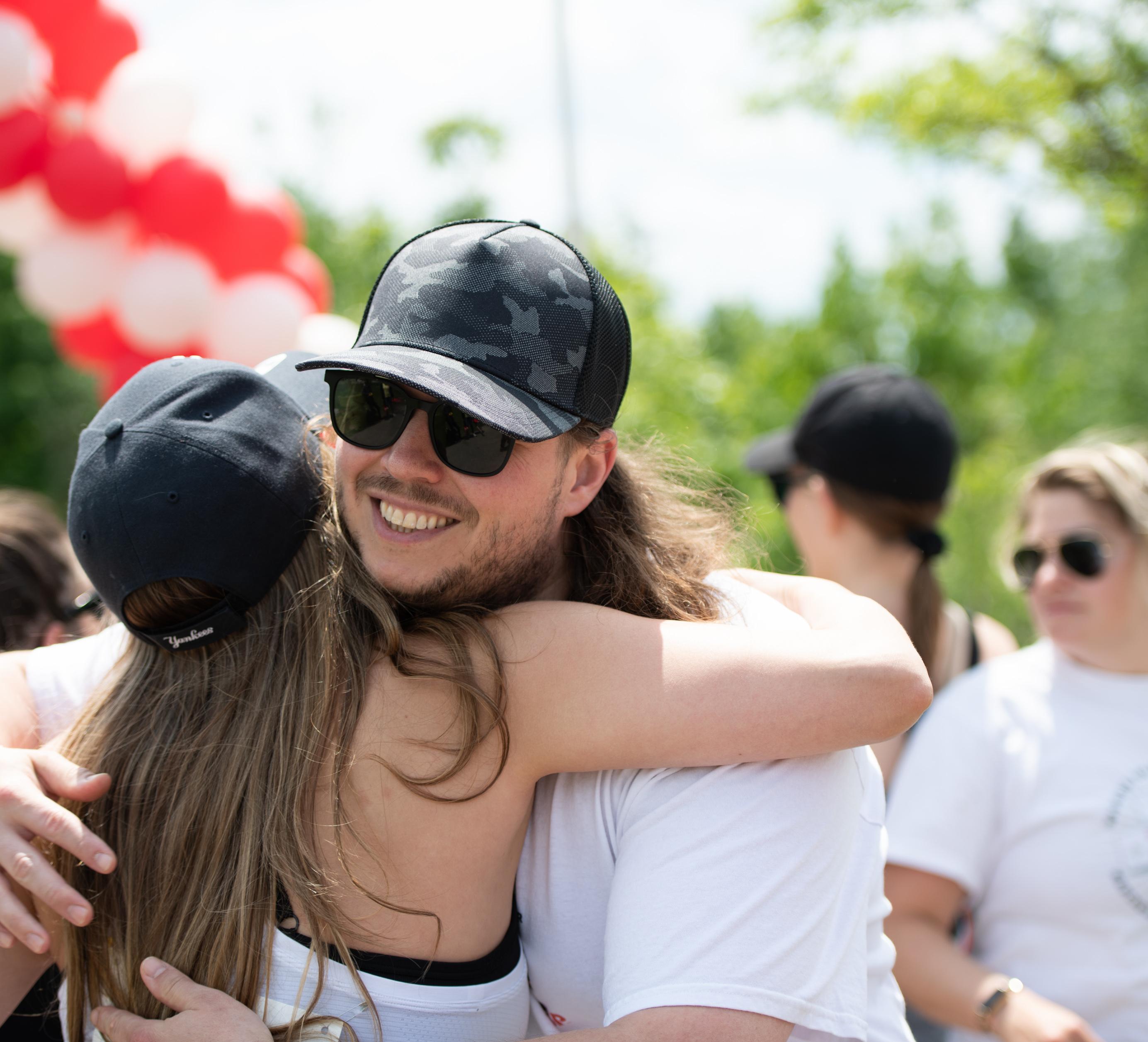 Two people with long hair and ball caps on embrace and smile