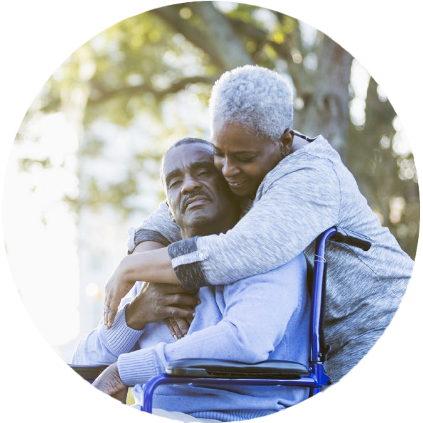 A woman with short grey hair hugs a man in a wheelchair from behind, resting her face on his lovingly.