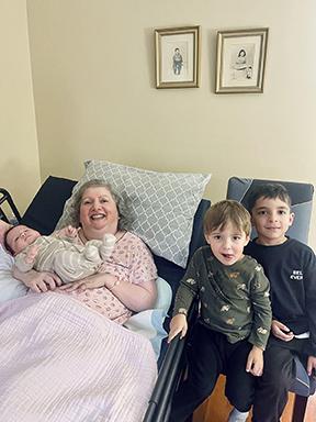 Carm est allongée dans son lit. Elle porte un pyjama rose et tient son petit-fils dans ses bras. Un autre de ses petits-fils et son petit-neveu sont assis à côté du lit.