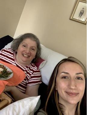 Carm est allongée dans son lit. Elle porte un t-shirt à rayures roses et tient une assiette de nourriture. Stephanie est à l’avant-plan et elle sourit.