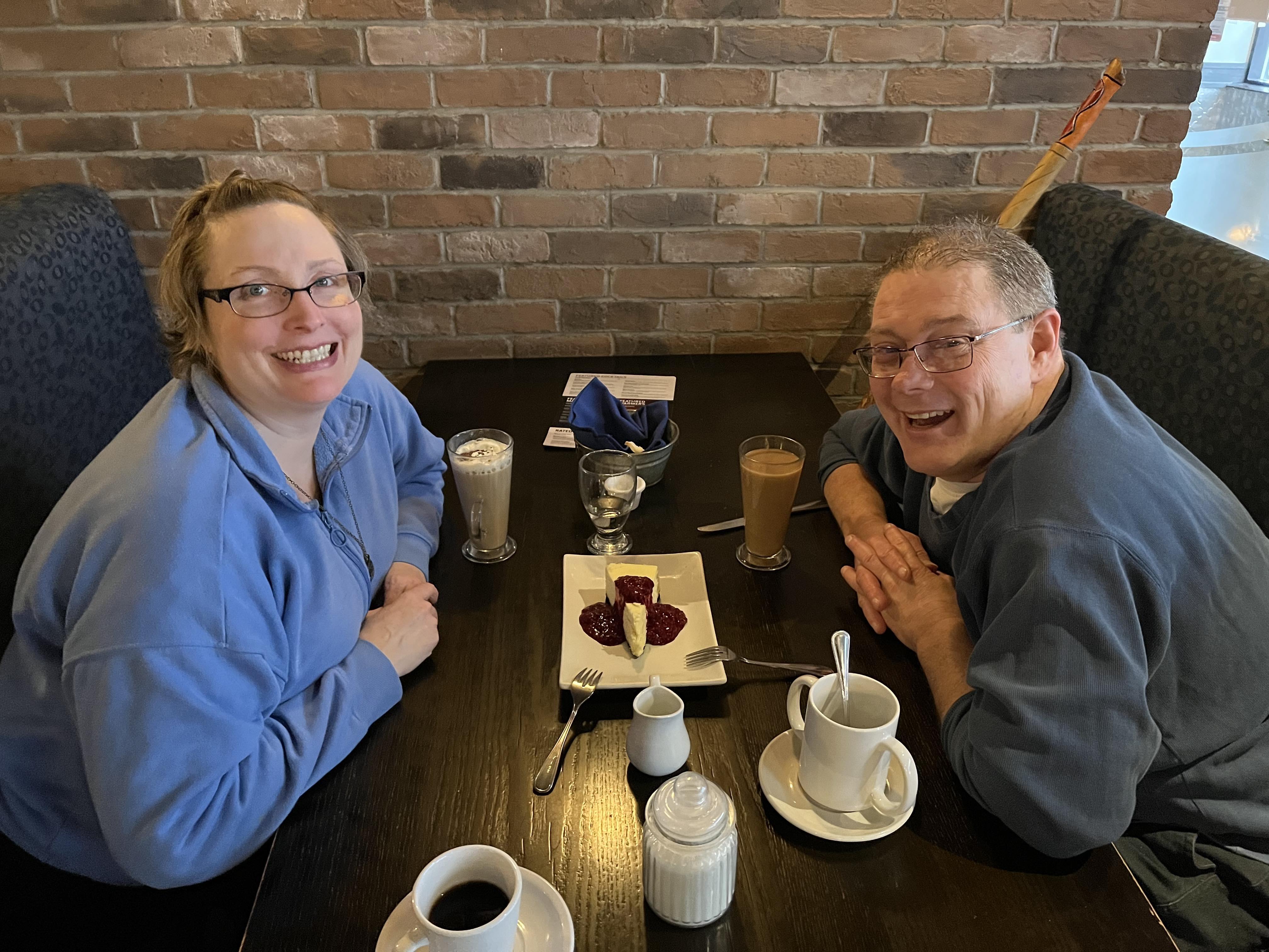 Yolanda porte un chandail bleu pâle, et James, un chandail gris foncé. Ils sont dans un restaurant, assis à une table sur laquelle un dessert et des boissons chaudes ont été déposés.