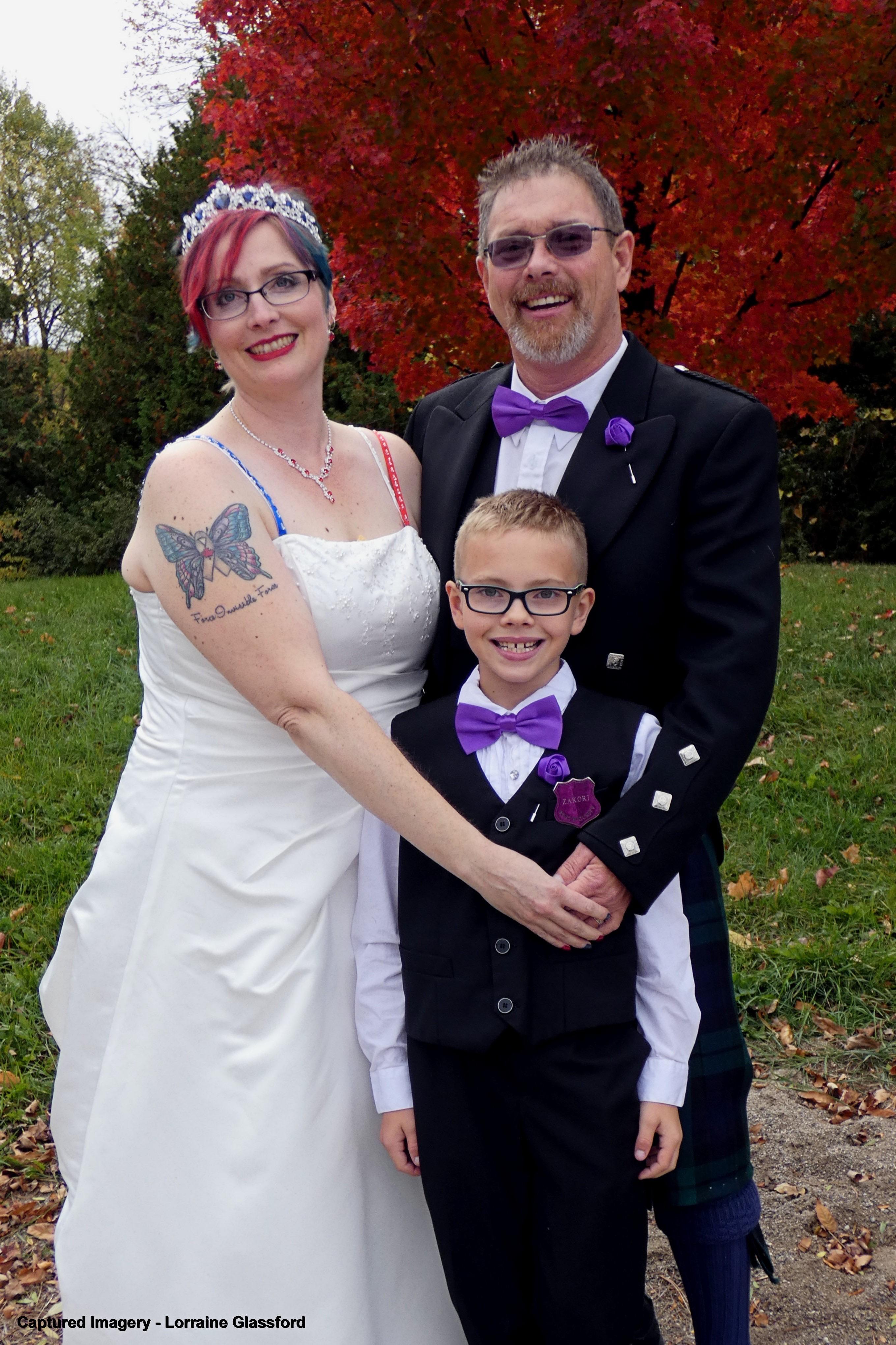 Yolanda, wearing a white wedding dress and a tiara, James wearing a suit and a purple bow tie, and their son, wearing a suit and purple bow tie, standing outside.