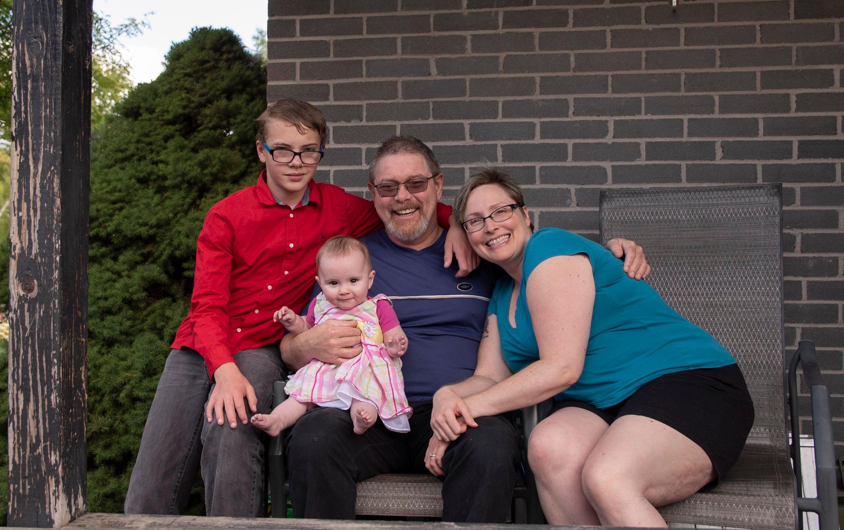 James, wearing a dark blue shirt, Yolanda, wearing a teal shirt, their son wearing a red button up shirt, and their daughter, wearing a pink striped dress, sitting outside.