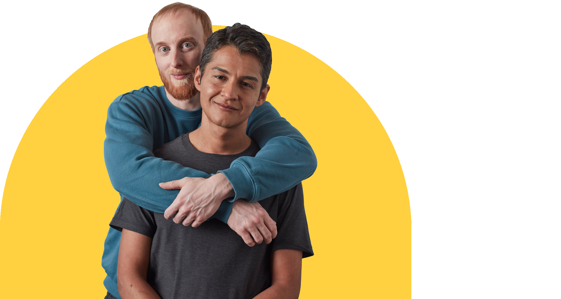 A man with short red hair and a beard, wearing a long sleeve blue shirt hugs a man with short brown hair in a tshirt