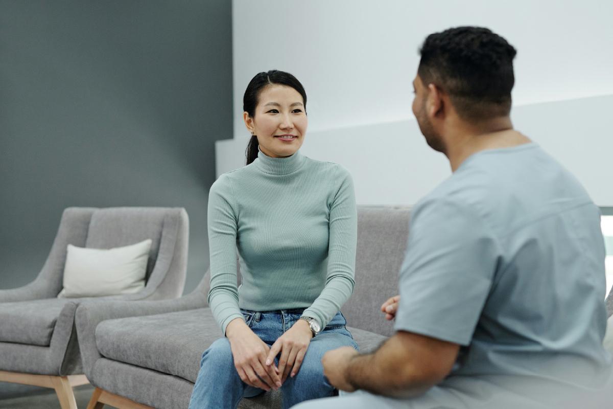 A woman sits on a couch facing a man who is dressed in healthcare scrubs.