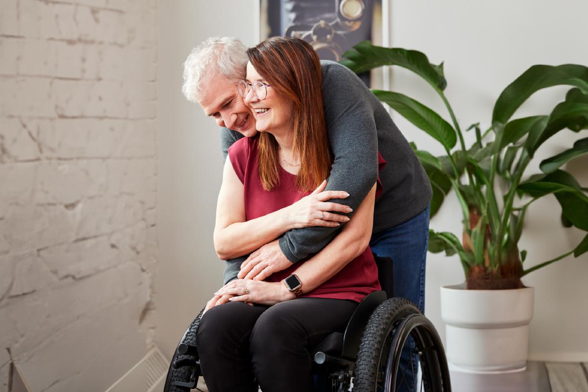 A woman in a wheelchair is being hugged from behind by a man.