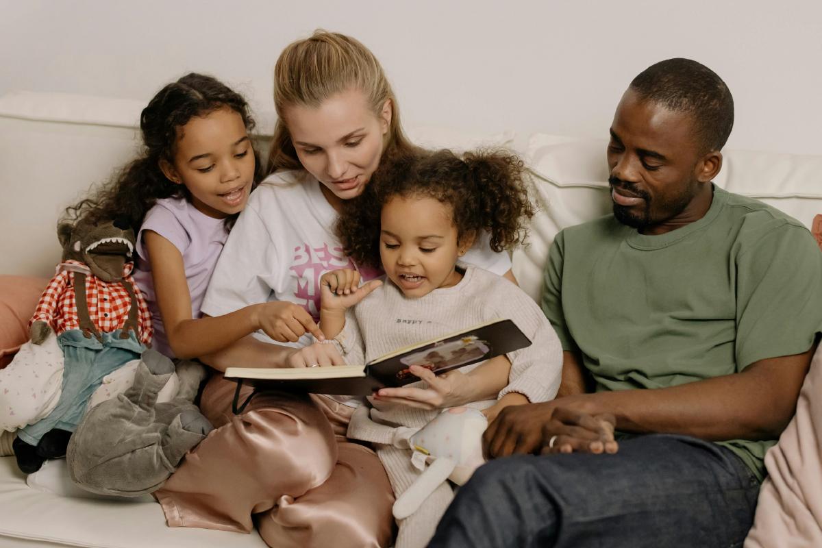 A family of four sits on the couch reading a picture book together.